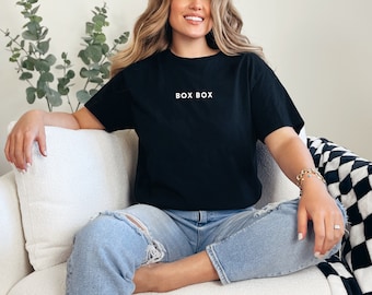 May include: A woman wearing a black t-shirt with the text "BOX BOX" in white lettering, blue jeans, and a gold bracelet. She is sitting on a white sofa.