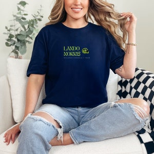 May include: A navy blue t-shirt with a green and white graphic of a race car driver's helmet and the text "Lando Norris McLaren Formula 1 Team".