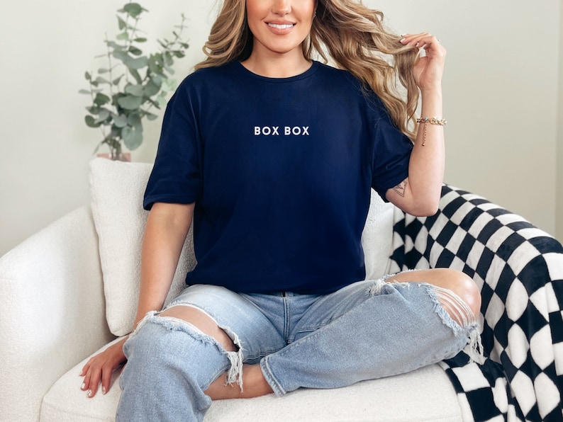 May include: A woman wearing a navy blue t-shirt with the text "BOX BOX" in white lettering. She is sitting on a white couch with a black and white checkered throw blanket.