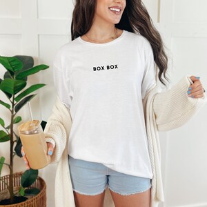 May include: A white t-shirt with the text "BOX BOX" printed on the front. The person is wearing denim shorts and a cream cardigan.