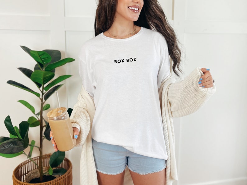 May include: A white t-shirt with the text "BOX BOX" printed on the front. The person is wearing denim shorts and a cream cardigan.