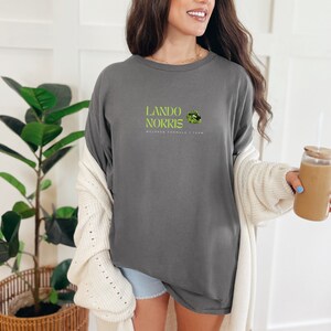 May include: A grey long-sleeved t-shirt with the text "Lando Norris McLaren Formula 1 Team" and a green and black graphic of a lip print.