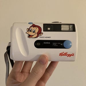 May include: A white Konica 35mm film camera featuring a Choco Monkey graphic and Kellogg's logo. The camera has a black lens housing with the text Konica Lens Japan f=33mm, a blue shutter button, a built-in flash, and a grey wrist strap attached.