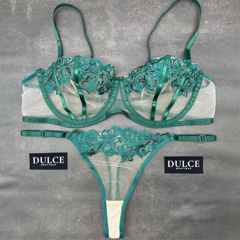 May include: A two-piece lingerie set in emerald green featuring a sheer mesh bra with underwire and matching thong. Both pieces are adorned with intricate floral embroidery and satin straps. Two black labels with the white text DULCE BOUTIQUE.