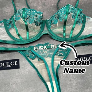 May include: An emerald green lingerie set consisting of a sheer underwire bra with floral embroidery and matching thong underwear. The thong features silver-toned metal lettering that spells out FUCK ME, with a graphic arrow pointing to it.