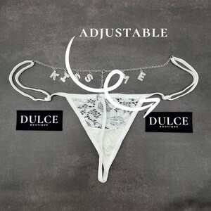 Puede incluir: Lingerie de encaje blanco con una cadena ajustable que presenta la palabra "KISS ME" en pedrer&iacute;a. La lencer&iacute;a tiene un delicado patr&oacute;n floral y est&aacute; etiquetada como "Dulce Boutique".