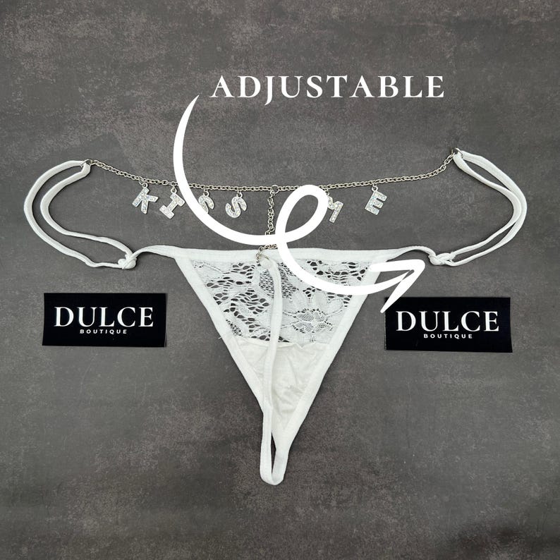 Puede incluir: Lingerie de encaje blanco con una cadena ajustable que presenta la palabra "KISS ME" en pedrer&iacute;a. La lencer&iacute;a tiene un delicado patr&oacute;n floral y est&aacute; etiquetada como "Dulce Boutique".
