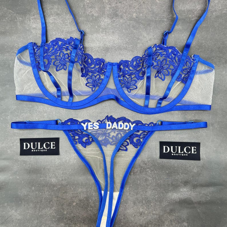 May include: A royal blue lingerie set consisting of a sheer underwire bra and a matching thong. The bra features floral embroidery and thin straps. The thong includes a silver-toned metal charm spelling out the text YES DADDY.