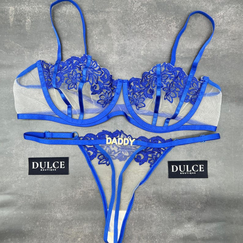 May include: A royal blue lingerie set consisting of an underwire bra and a thong. The bra features sheer mesh panels with blue floral embroidery and thin straps. The matching thong includes blue floral embroidery and a gold-toned metal charm that reads DADDY.
