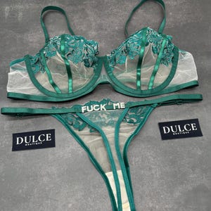 May include: A dark emerald green lingerie set consisting of a sheer underwire bra and a matching thong. The bra features floral embroidery and satin straps. The thong includes a silver-toned metal charm with the text FUCK ME. Two black tags read DULCE BOUTIQUE.
