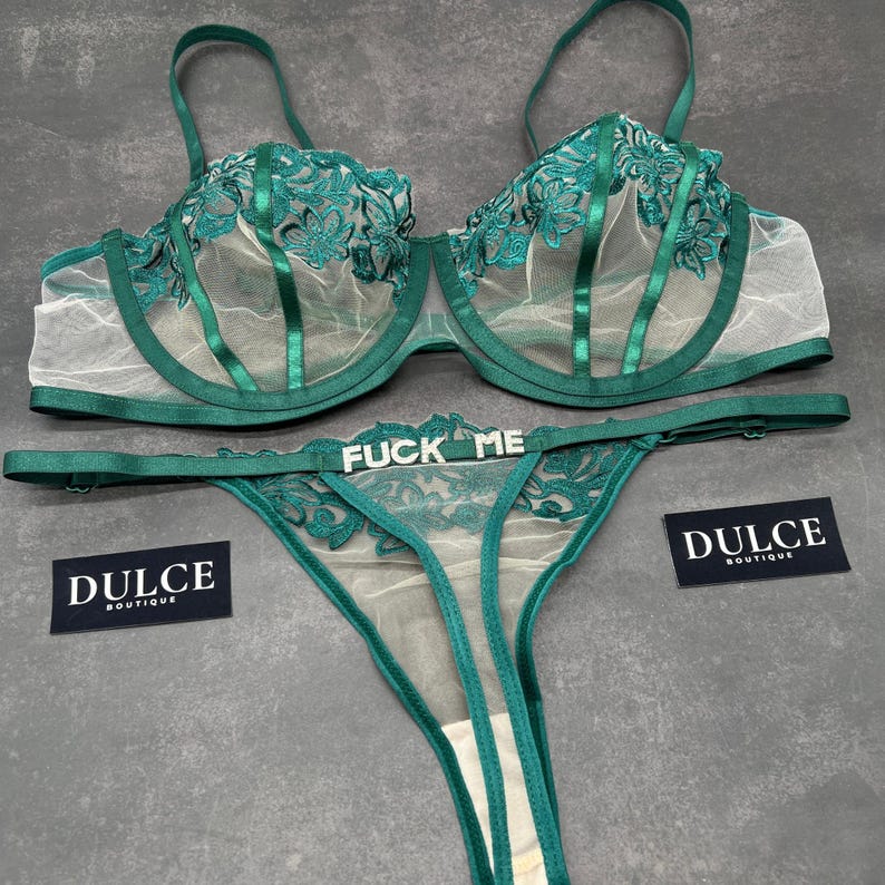May include: A dark emerald green lingerie set consisting of a sheer underwire bra and a matching thong. The bra features floral embroidery and satin straps. The thong includes a silver-toned metal charm with the text FUCK ME. Two black tags read DULCE BOUTIQUE.
