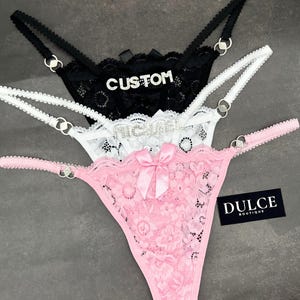 Könnte beinhalten: Drei Paar Spitzenunterwäsche in Pink, Weiß und Schwarz. Das rosafarbene Paar hat eine Schleife aus Satin und den Text "DULCE BOUTIQUE". Das weiße Paar hat den Text "MICHAEL" und das schwarze Paar hat den Text "CUSTOM".