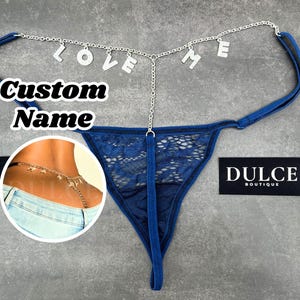 Op de afbeelding: Een blauwe kanten string met een zilverkleurige kettingtailleband en bungelende letterbedels die LOVE ME spellen. Een cirkelvormige afbeelding toont de ketting gedragen rond de taille boven een blauwe spijkerbroek. Tekst op de afbeelding: Custom Name en DULCE BOUTIQUE.
