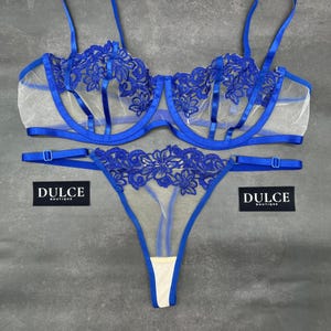 May include: A blue lingerie set consisting of a bra and a thong, both featuring sheer mesh fabric and intricate blue floral embroidery. The bra has adjustable straps and underwire support. Two black tags with the white text DULCE BOUTIQUE are visible.