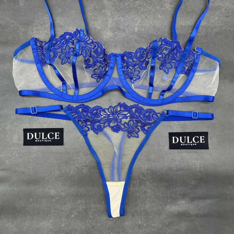 May include: A blue lingerie set consisting of a bra and a thong, both featuring sheer mesh fabric and intricate blue floral embroidery. The bra has adjustable straps and underwire support. Two black tags with the white text DULCE BOUTIQUE are visible.