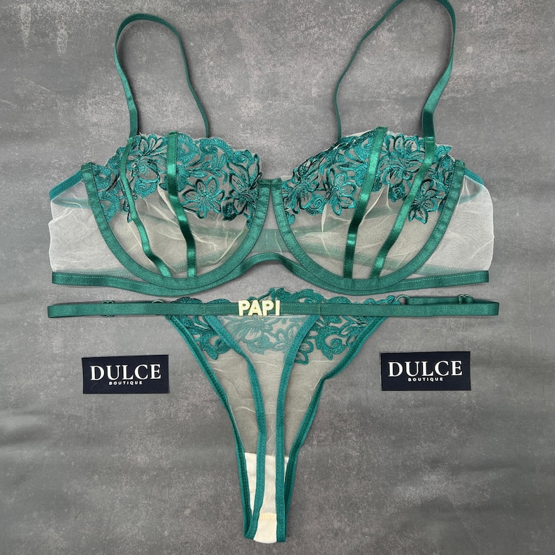 May include: A dark emerald green lingerie set consisting of a sheer underwire bra and a matching thong. The bra features floral embroidery and thin straps. The thong includes a gold-toned metal plaque with the text PAPI. Two black labels read DULCE BOUTIQUE.