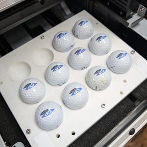 May include: A white tray with ten golf balls inside. The golf balls are white with a blue and white logo that says "CP". The tray is in a machine that appears to be a golf ball printing machine.