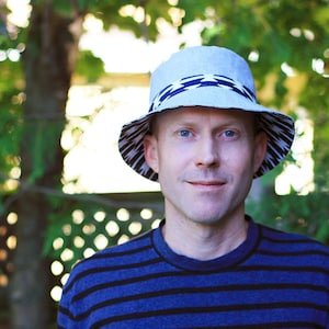 以下が含まれることがあります： 青と黒のストライプのシャツを着た男性が、白黒の幾何学模様のグレーのバケットハットをかぶっています