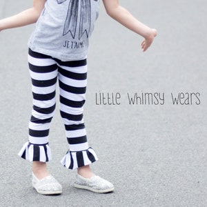 Puede incluir: Una niña pequeña con una camiseta gris con un gráfico de lazo blanco y negro y el texto "Je T'aime". También lleva pantalones negros y blancos a rayas con un detalle de volantes en la parte inferior y zapatos blancos.