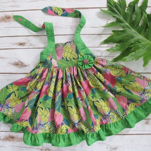 May include: A green and pink floral print dress with a green ruffle trim and straps. The dress has a single button closure at the back of the neck.