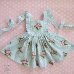 Könnte beinhalten: Ein hellblaues Kleid mit floralem Muster, quadratischem Ausschnitt und Rüschenträgern. Das Kleid hat einen weiten Rock und ist mit rosa Rosen und grünen Blättern verziert. Das Kleid hat lange Bindebänder und den Text "tie dye Diva sewing patterns".