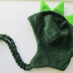 May include: Green fleece hat with a green and lime green dinosaur-like design. The hat has a long, braided tail.