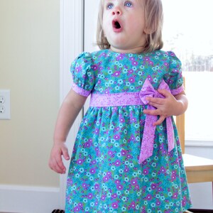 May include: A young girl wearing a blue and purple floral dress with a purple bow. The dress has a puffed sleeve and a gathered skirt.