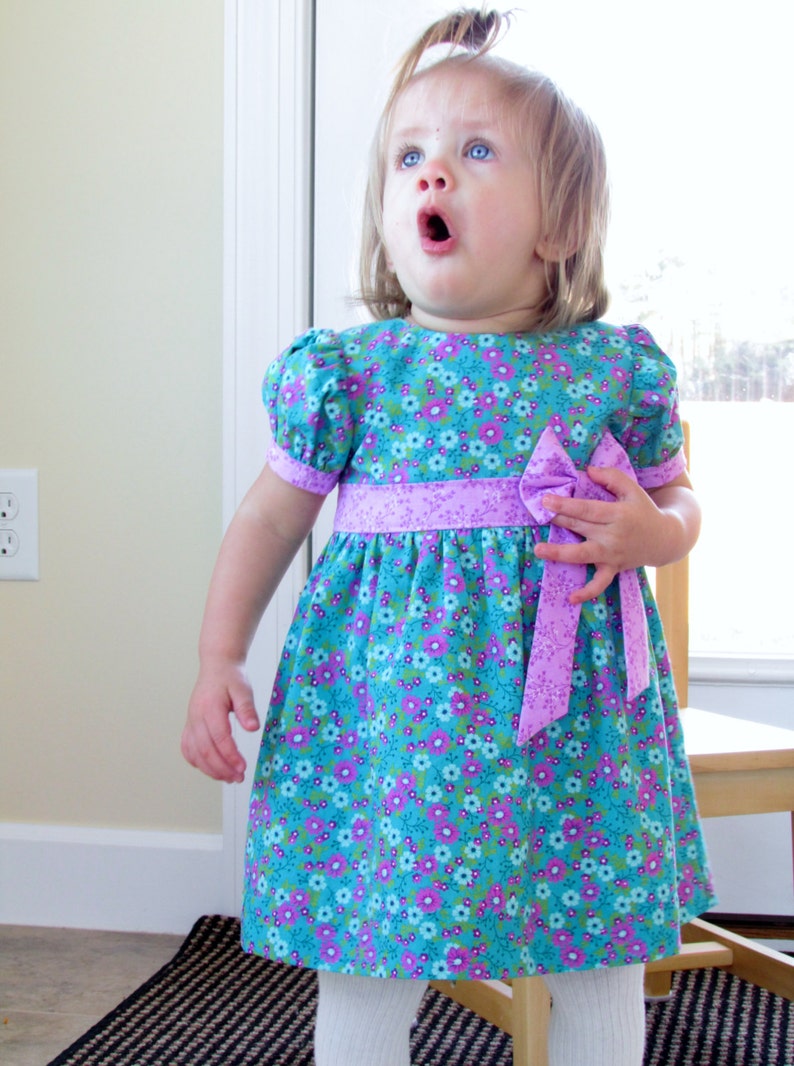 May include: A young girl wearing a blue and purple floral dress with a purple bow. The dress has a puffed sleeve and a gathered skirt.