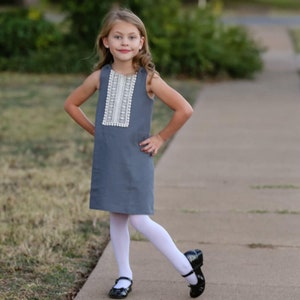 May include: A sleeveless, slate-gray dress with a white lace and pom-pom trim detail on the bodice. The dress is paired with white tights and black Mary Jane shoes. The girl is standing on a sidewalk.