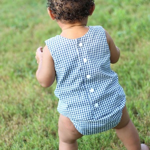 May include: A toddler wearing a blue and white gingham romper with white buttons down the back. The toddler is standing on a grassy lawn.