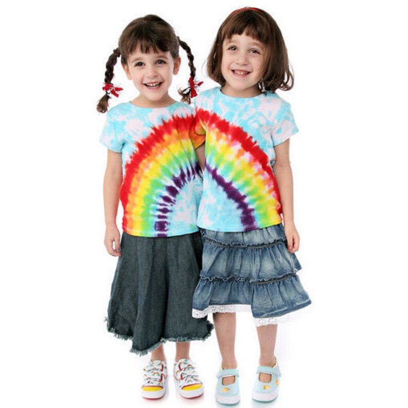 May include: Two young girls wearing tie-dye shirts and denim skirts. The shirts have a rainbow design. The girls are smiling and looking at the camera.