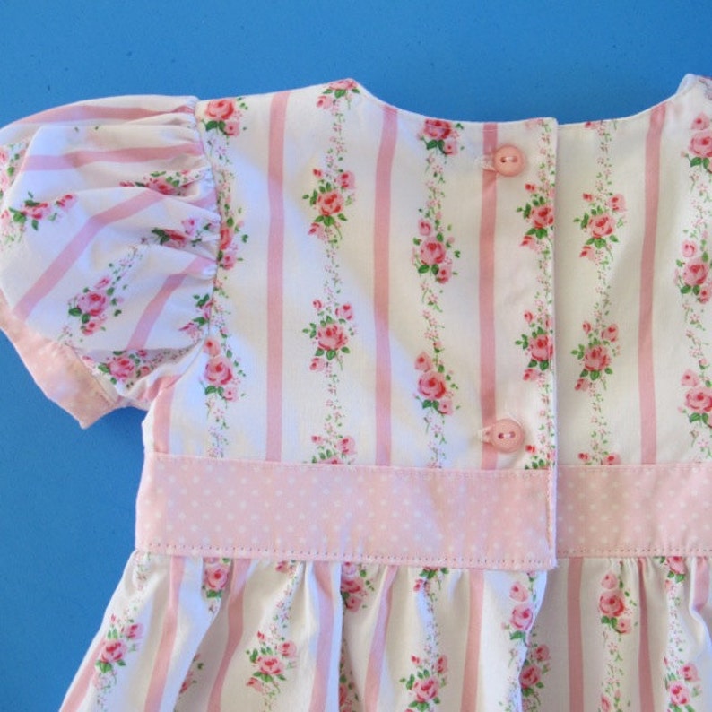 May include: A white and pink striped dress with a pink floral pattern and a pink polka dot waistband. The dress has a button closure in the back.
