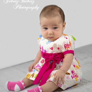May include: A baby girl wearing a white dress with a pink bow and colorful cartoon animals and flowers. She is sitting on a white wooden floor and wearing pink socks with white polka dots.