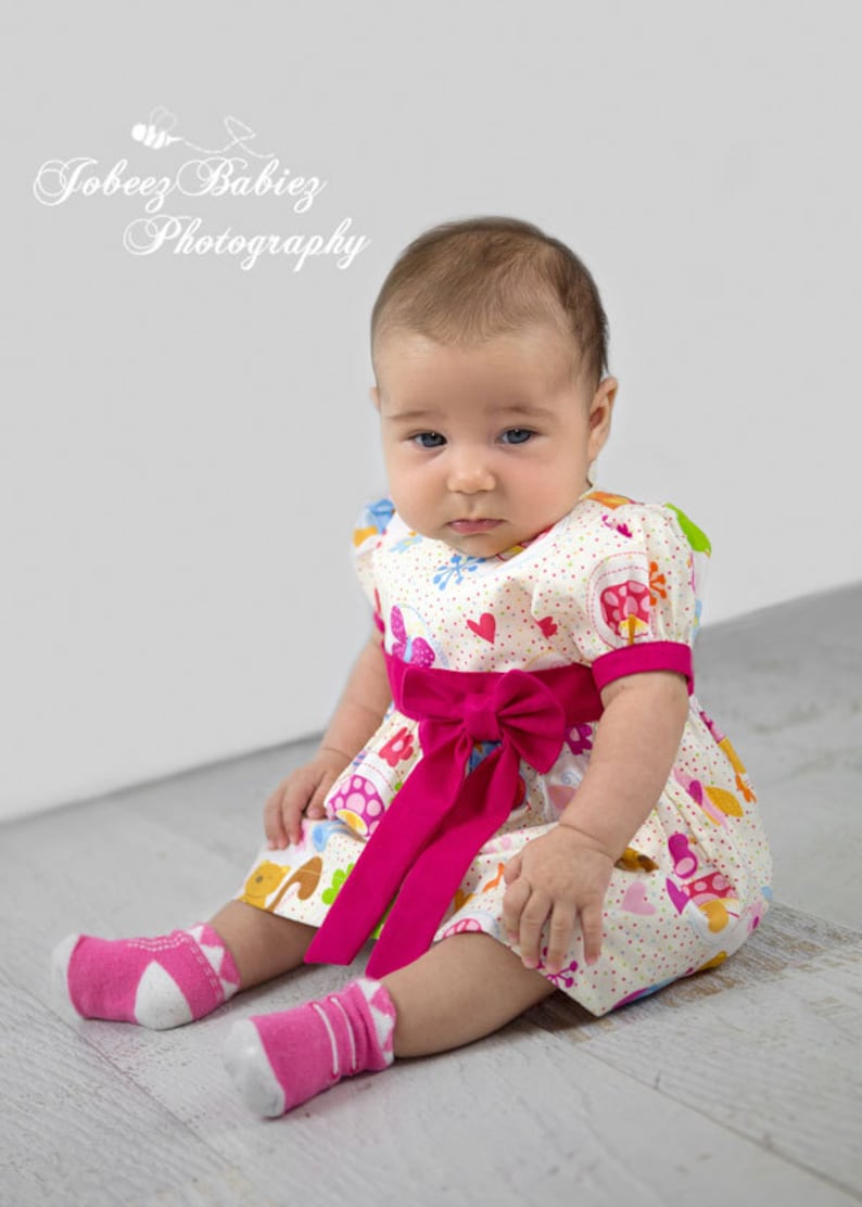 May include: A baby girl wearing a white dress with a pink bow and colorful cartoon animals and flowers. She is sitting on a white wooden floor and wearing pink socks with white polka dots.