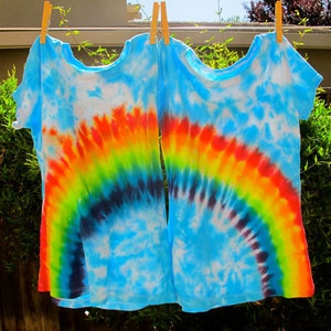May include: Two tie-dye t-shirts with a rainbow design. The shirts are blue with a rainbow arc in shades of yellow, orange, red, green, and blue.