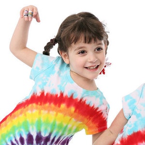 May include: A young girl with brown hair in pigtails is wearing a tie-dye shirt with a rainbow pattern. She is smiling and has her arm raised.