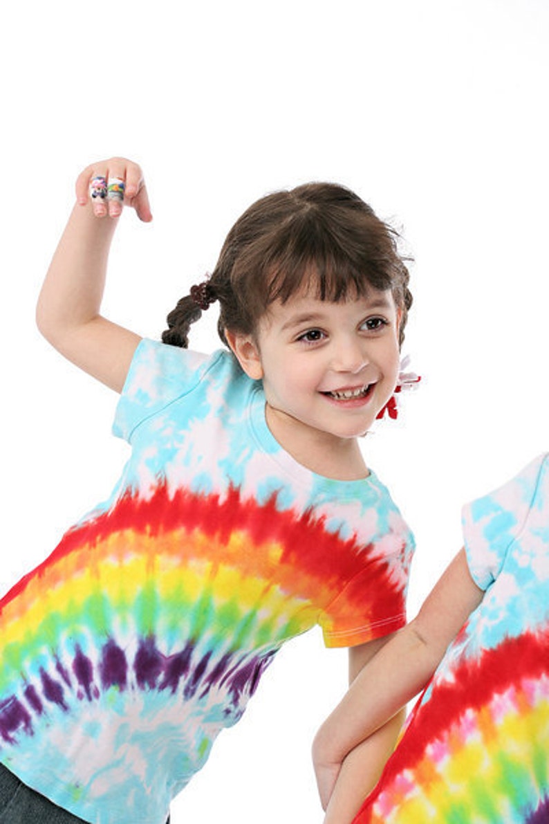 May include: A young girl with brown hair in pigtails is wearing a tie-dye shirt with a rainbow pattern. She is smiling and has her arm raised.