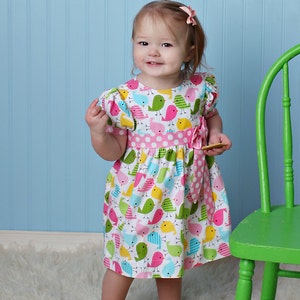 May include: A young girl wearing a white dress with a pink polka dot sash and a colorful bird print. The dress has short sleeves and a bow at the waist. The girl is standing on a white fluffy rug in front of a green chair.