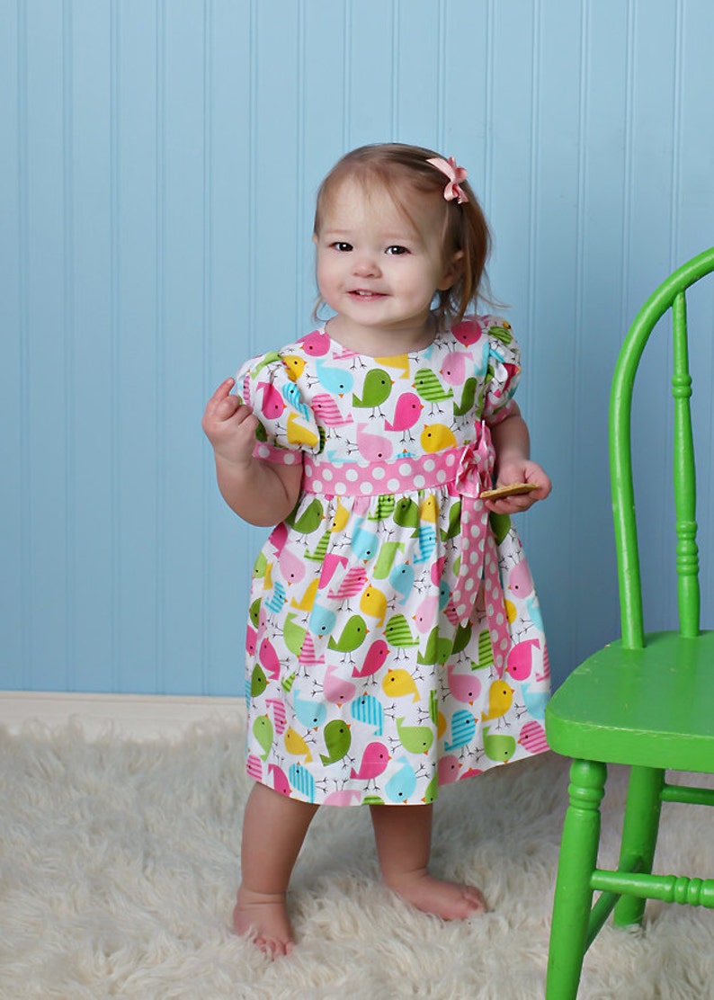 May include: A young girl wearing a white dress with a pink polka dot sash and a colorful bird print. The dress has short sleeves and a bow at the waist. The girl is standing on a white fluffy rug in front of a green chair.