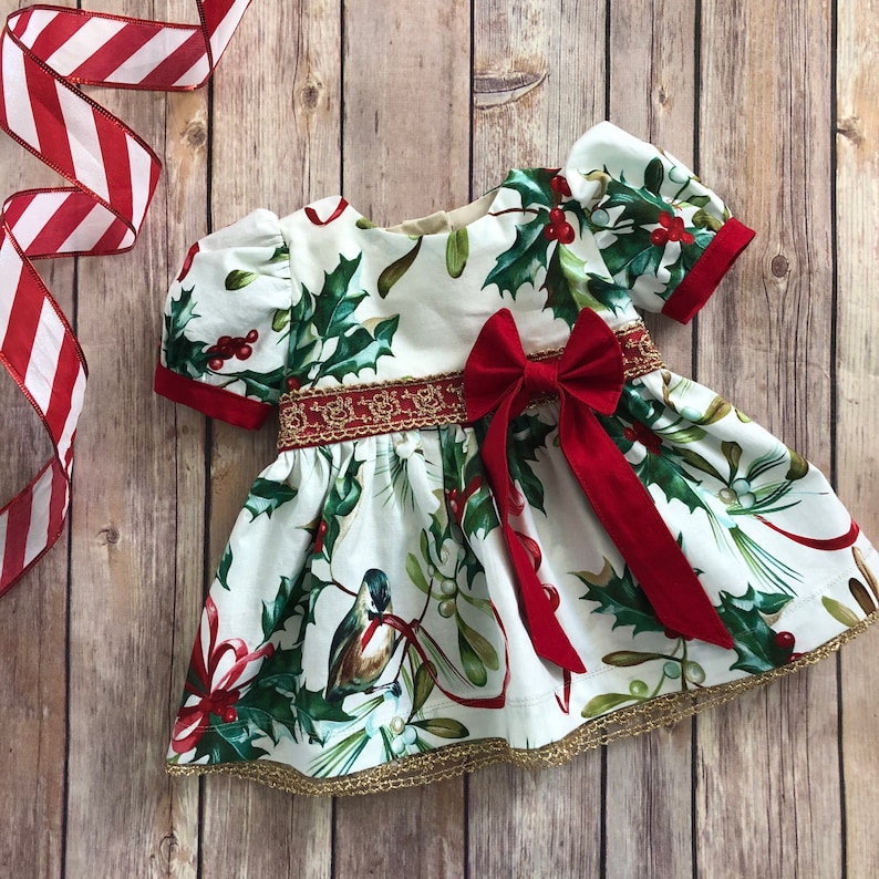 May include: A white dress with a red bow and a holly and bird print. The dress has a gold trim and a red trim around the sleeves.