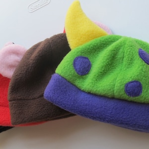 May include: Four fleece hats with different animal ears. The hats are in various colors, including black, red, brown, green, yellow, and purple. The hats have different ear shapes, including cat ears, devil horns, pig ears, and horns.