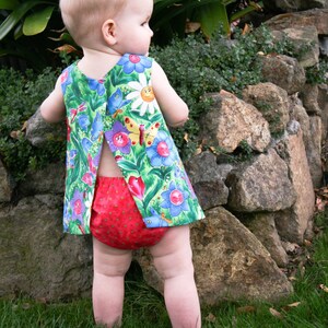 May include: A baby wearing a colorful floral print dress and red diaper cover stands on grass. The dress features a green background with blue, purple, and yellow flowers and butterflies. The baby is standing near a stone wall.