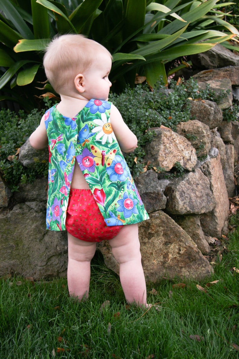 May include: A baby wearing a colorful floral print dress and red diaper cover stands on grass. The dress features a green background with blue, purple, and yellow flowers and butterflies. The baby is standing near a stone wall.
