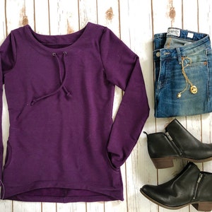 Könnte beinhalten: Ein lilafarbenes Langarmshirt mit einem Kordelzug am Ausschnitt und Seitentaschen. Das Shirt wird mit einer blauen Jeans und schwarzen Stiefeletten kombiniert.