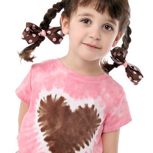 May include: A young girl with brown hair wearing a pink tie-dye shirt with a brown heart design. She has two brown bows with white polka dots in her hair.