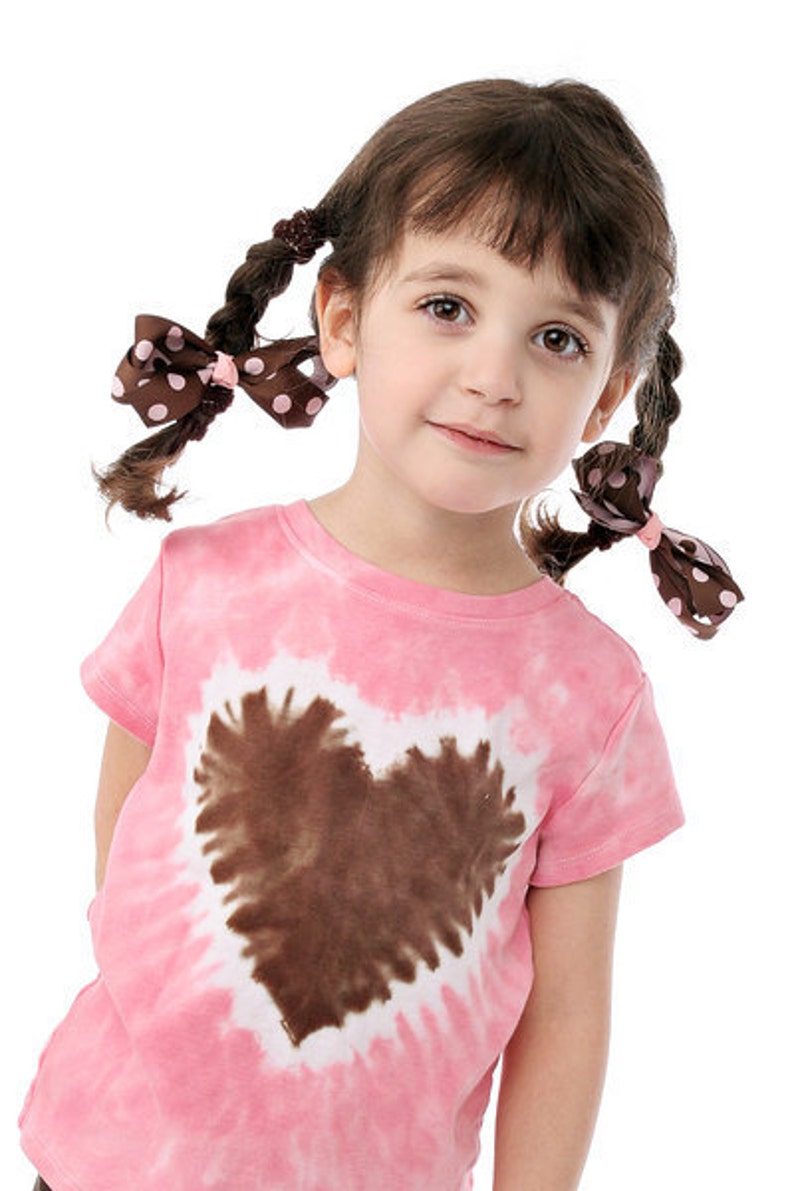 May include: A young girl with brown hair wearing a pink tie-dye shirt with a brown heart design. She has two brown bows with white polka dots in her hair.