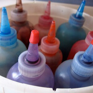 May include: A close-up of a white bucket filled with various colored bottles of dye. The bottles have pointed tops and are arranged in a circle. The colors include blue, red, purple, and orange.