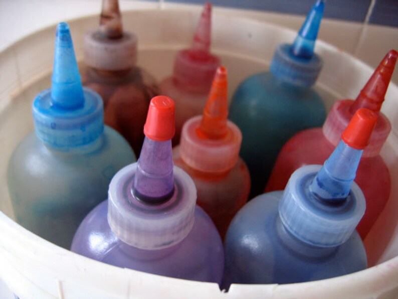 May include: A close-up of a white bucket filled with various colored bottles of dye. The bottles have pointed tops and are arranged in a circle. The colors include blue, red, purple, and orange.