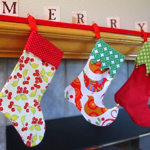 Puede incluir: Tres calcetines de Navidad colgados de una repisa de madera con la palabra "Merry" escrita en bloques de madera. Los calcetines están decorados con diferentes patrones, incluyendo lunares rojos y blancos, círculos verdes y blancos y lunares verdes y blancos.