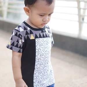 Puede incluir: Un niño pequeño con una camisa de patrón blanco y negro con un patrón geométrico gris y amarillo en las mangas. La camisa tiene un estampado gráfico blanco y negro en la parte delantera con el texto "GOON FC HI!" El niño lleva pantalones azules.